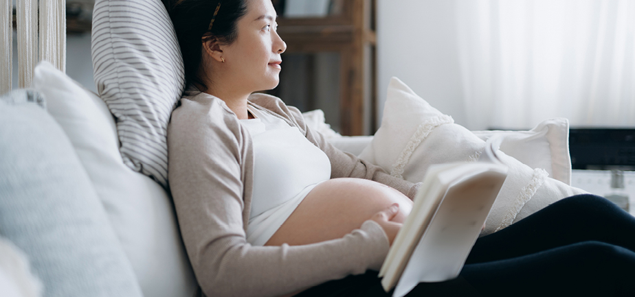 Asian pregnant woman relaxing at cozy home, looking thoughtful while reading a baby book on sofa.