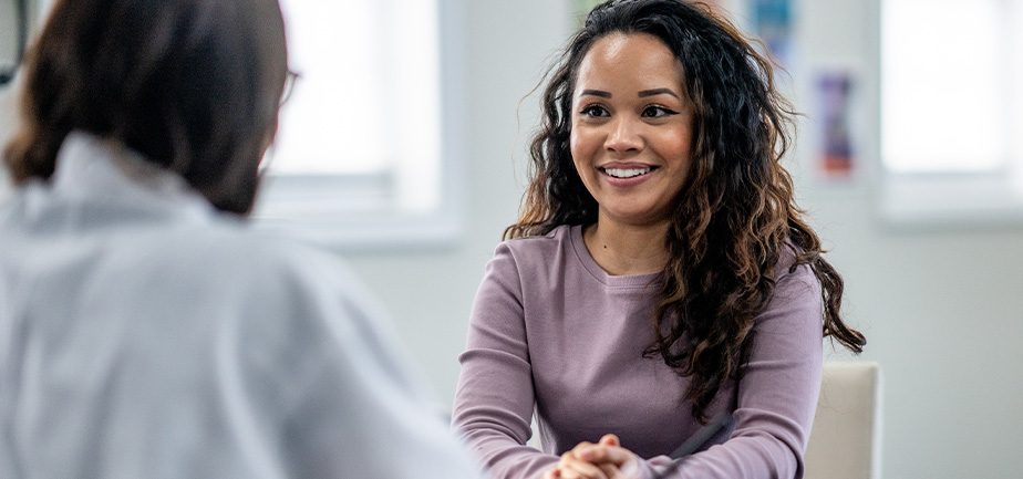 A young woman of mixed race, sits with her female doctor as they discuss her health concerns. The patient is dressed casually and has a smile on her face.