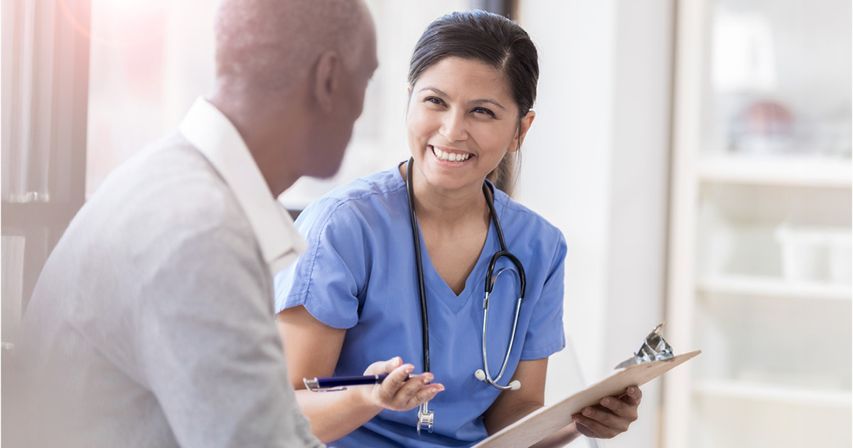 A smiling female doctor wears scrubs as she sits beside her unrecognizable senior male patient. She gestures as she holds a clipboard and shares his test results.