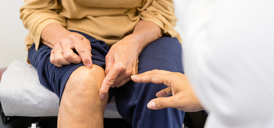 Senior adult woman, sitting on the examination table, shows doctor where her knee hurts.