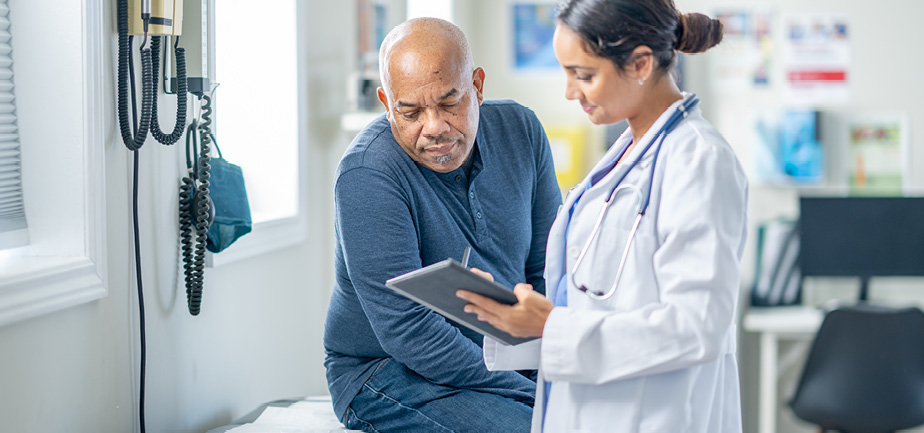 A senior African American gentleman sits up on an exam table during a routine check-up reviewing test results on a tablet with a female doctor. 
