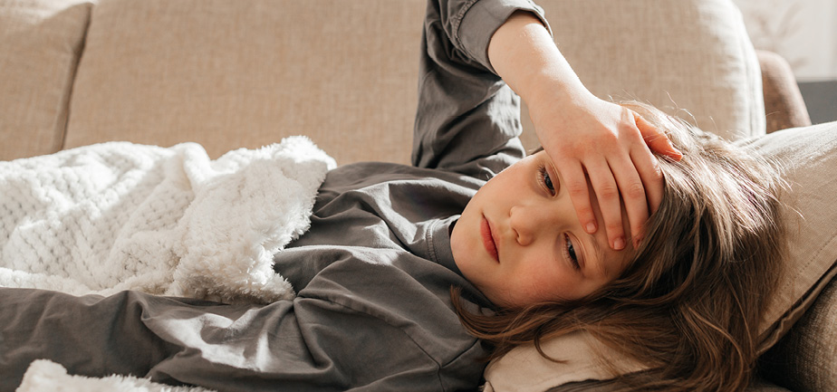 A sick dark-haired teenage girl lies on the sofa, covered with a blanket and put her hand on her forehead.