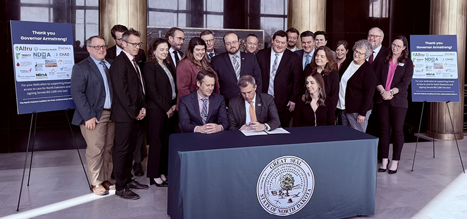 Large group of people gathered around a table where three people are sitting, with one man signing a document.