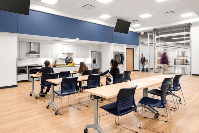 a classroom with a kitchen at the front and people sitting at rows of tables